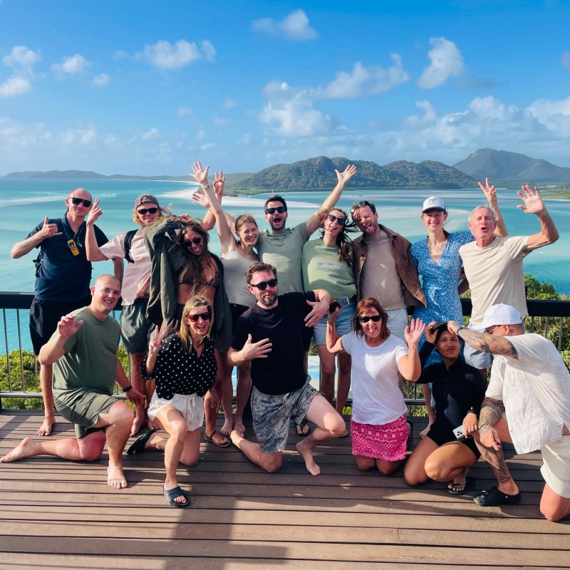 Group of people posing energetically on a deck with ocean and islands in the background.