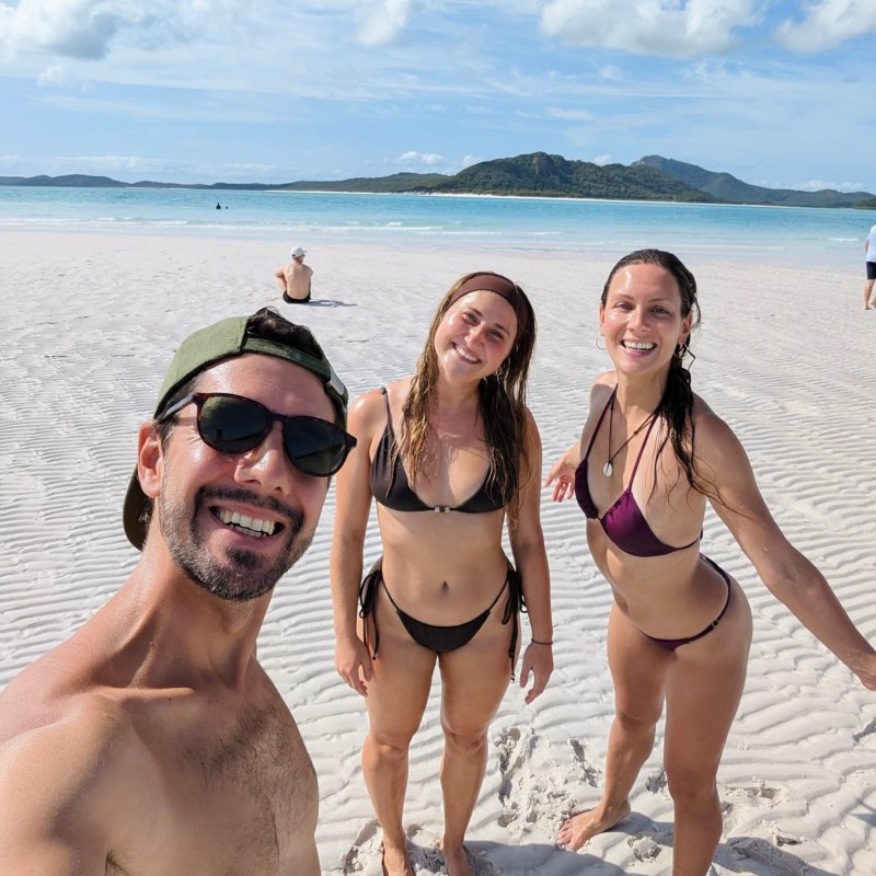 Three people smiling on a sandy beach with a mountain view.