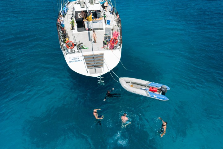 People swimming near a boat and a small dinghy in clear blue water.