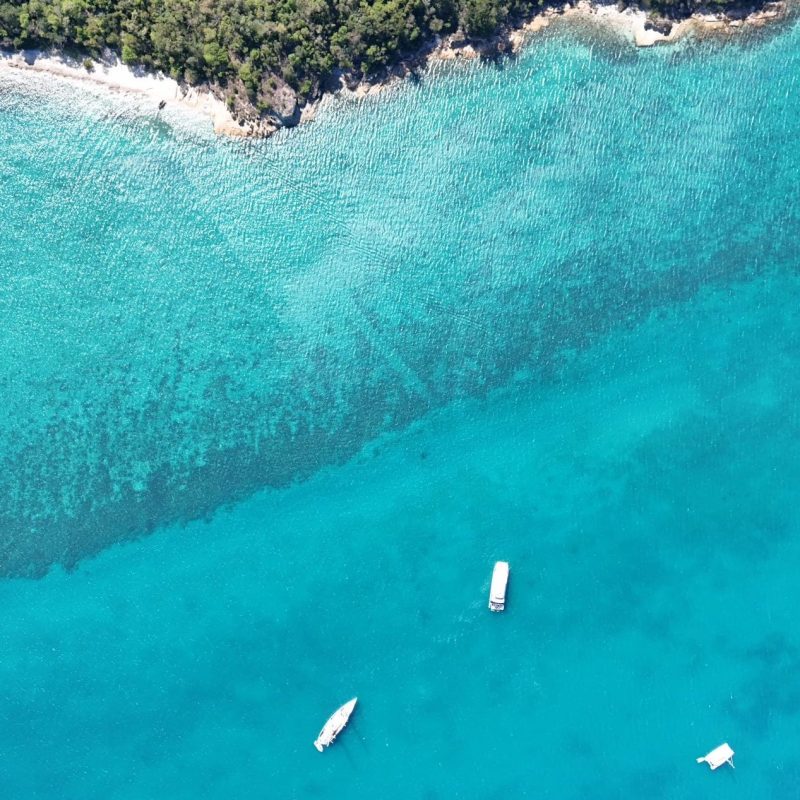 Aerial view of turquoise sea with several small boats and a rocky coastline.