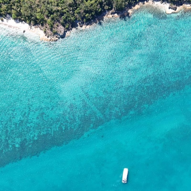 Aerial view of turquoise sea with several small boats and a rocky coastline.