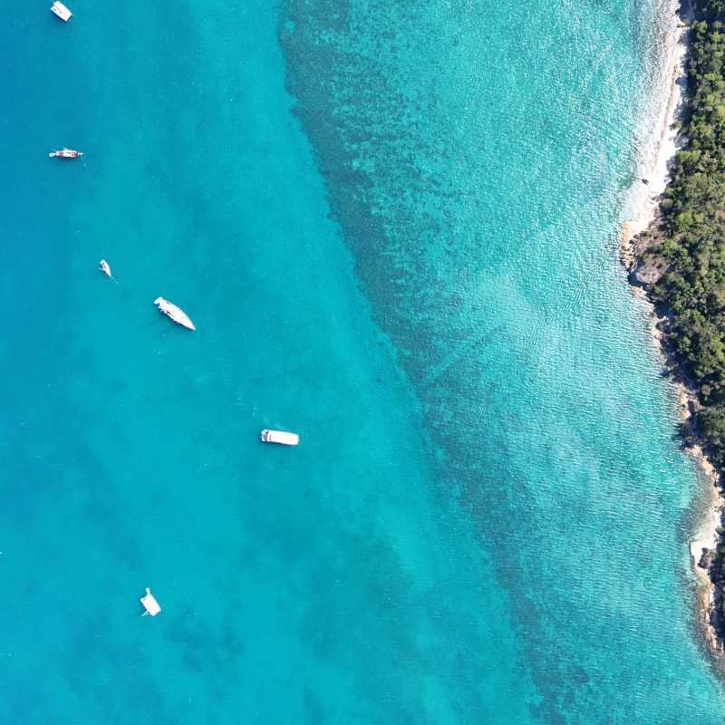 Aerial view of turquoise sea with several small boats and a rocky coastline.