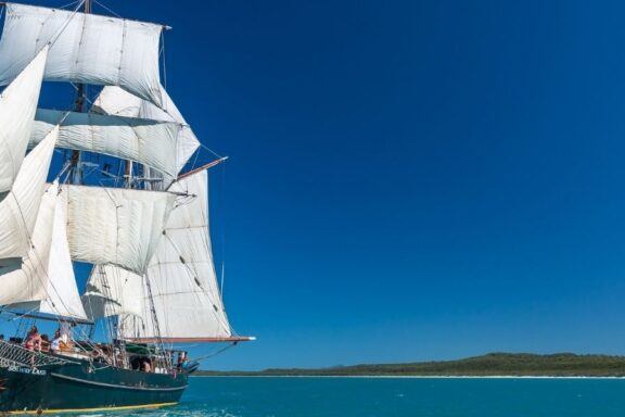 Tall ship with white sails at sea under clear blue sky, near coastline.