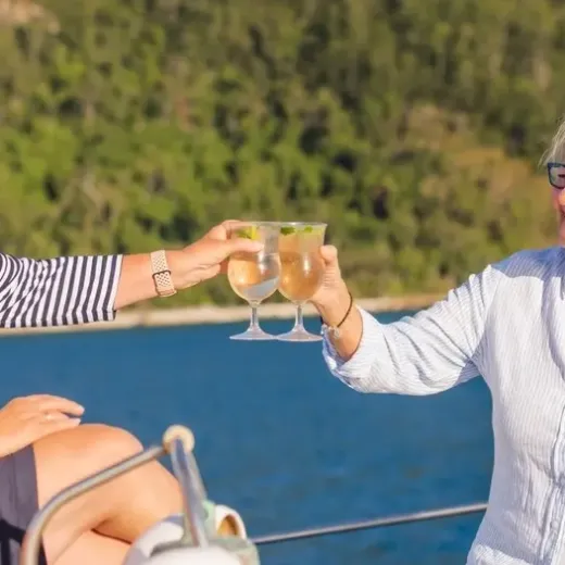 Two people toast with drinks on a boat, surrounded by water and trees.