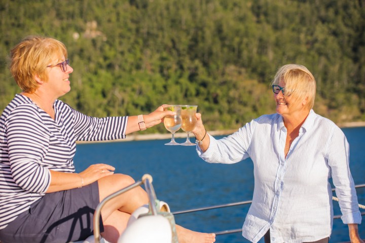 Two people on a boat clink glasses, smiling against a backdrop of water and forested hills.