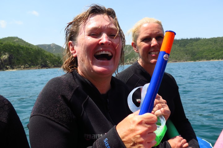 Two people in wetsuits, smiling on a boat with snorkeling gear, ocean and hills in the background.
