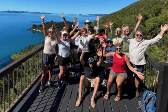 Group of people on a wooden deck by the ocean, all raising their hands in celebration.