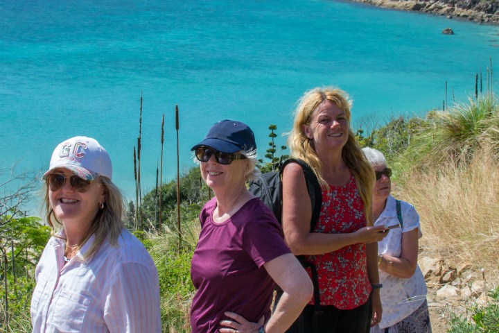Four women smiling on a coastal path with turquoise sea and cliffs in the background.