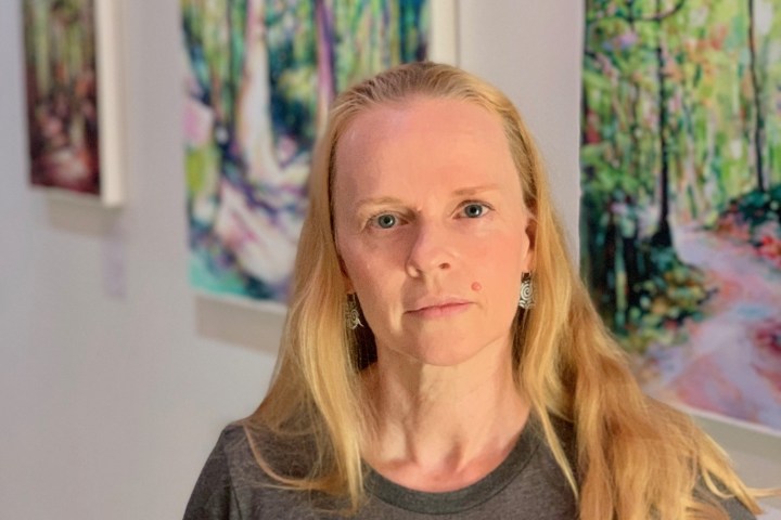 Woman with long blonde hair stands in front of colorful forest paintings in a gallery.