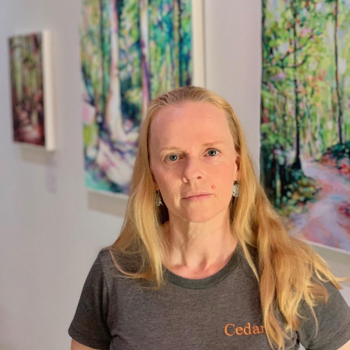 Woman with long blonde hair stands in front of colorful forest paintings in a gallery.
