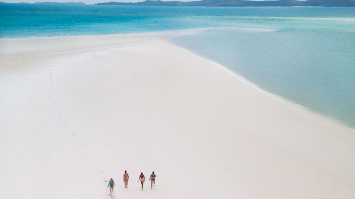 Four people walking on a vast white sandy beach with turquoise water under a blue sky.