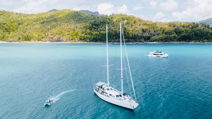 Sailboats on blue water near a forested coastline under a cloudy sky.